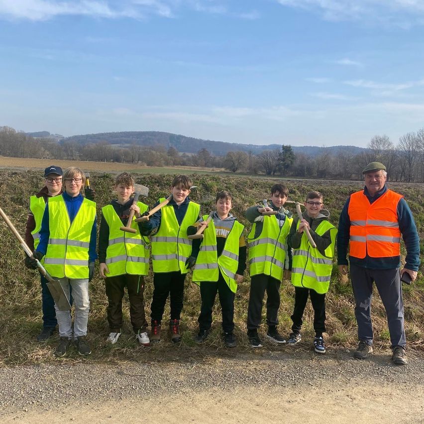 Eine Gruppe von Kindern und ein Mann stehen auf einem Pfad in einem Grasfeld, halten Schaufeln und tragen Warnwesten. Der Mann trägt eine orange Weste. Der Hintergrund zeigt einen Hügel mit Bäumen.