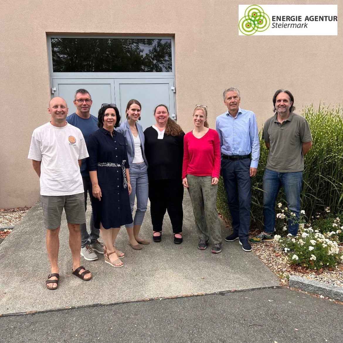 Eine Gruppe von Menschen, sowohl Männer als auch Frauen, stehen vor einem Gebäude und lächeln für ein Foto. Sie sind lässig gekleidet und stehen vor einer Wand mit einem Logo.