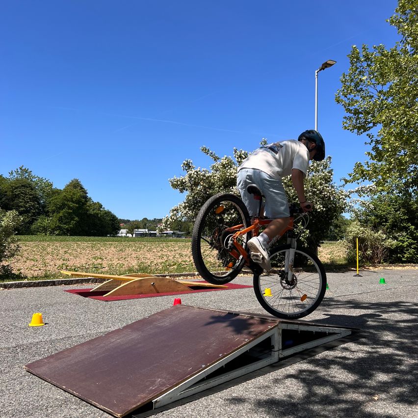 A cyclist wearing a helmet is airborne over a ramp in an outdoor park, surrounded by green trees and bushes.
