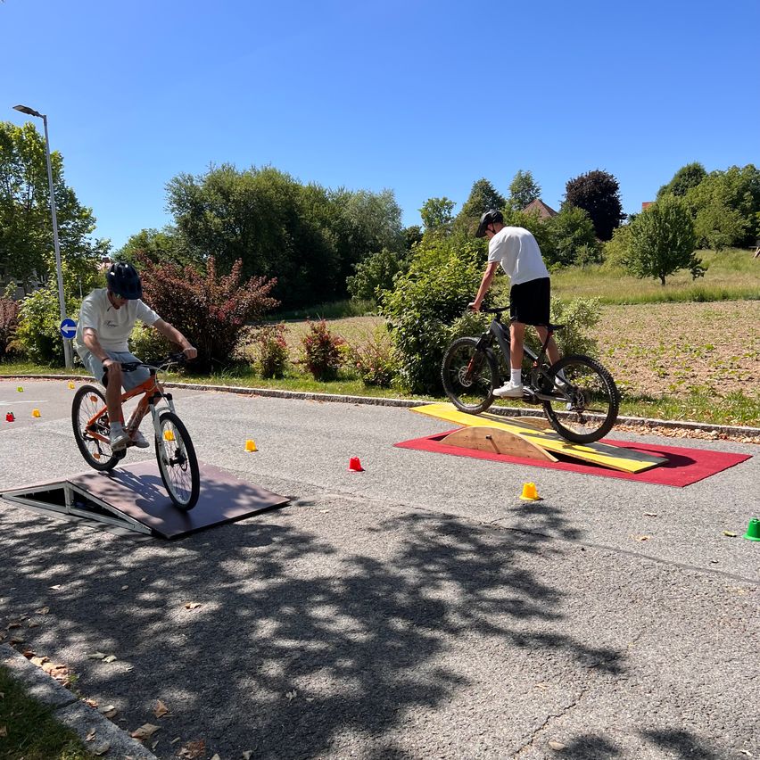 Two men are cycling on an outdoor track, navigating obstacles. One is on a black bike, the other on an orange one.