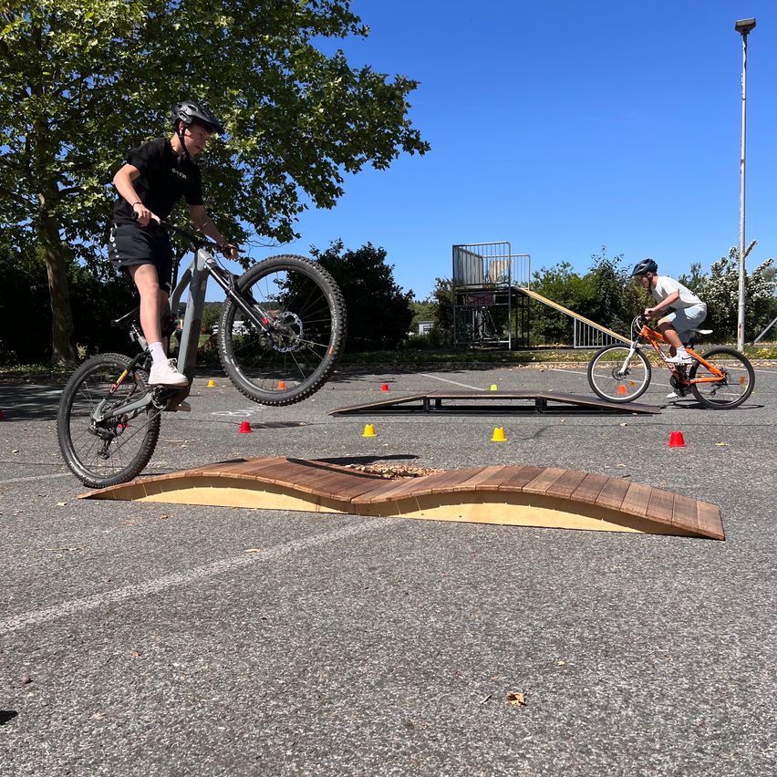 Two individuals ride bicycles on a paved area with a wooden ramp and orange cones. One rider approaches the ramp while the other navigates a sloped surface. Trees and a streetlight are visible in the background.
