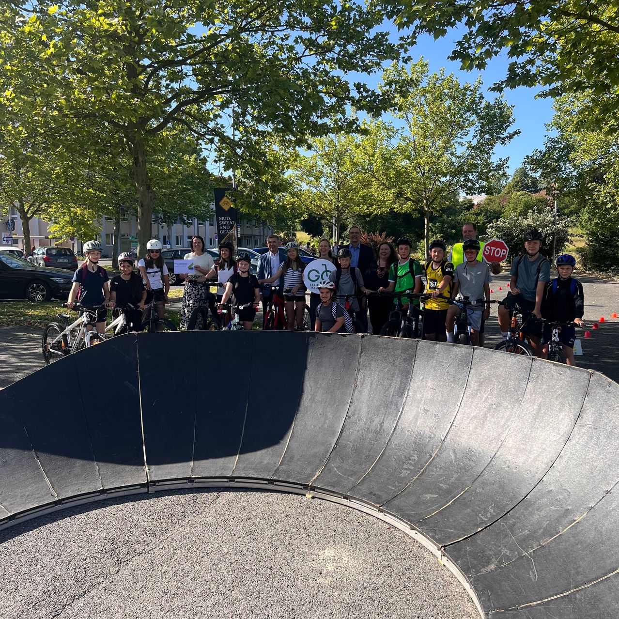 A group of young cyclists, some with helmets, gather with adults around a ramp, under trees with a stop sign and a building in the background.