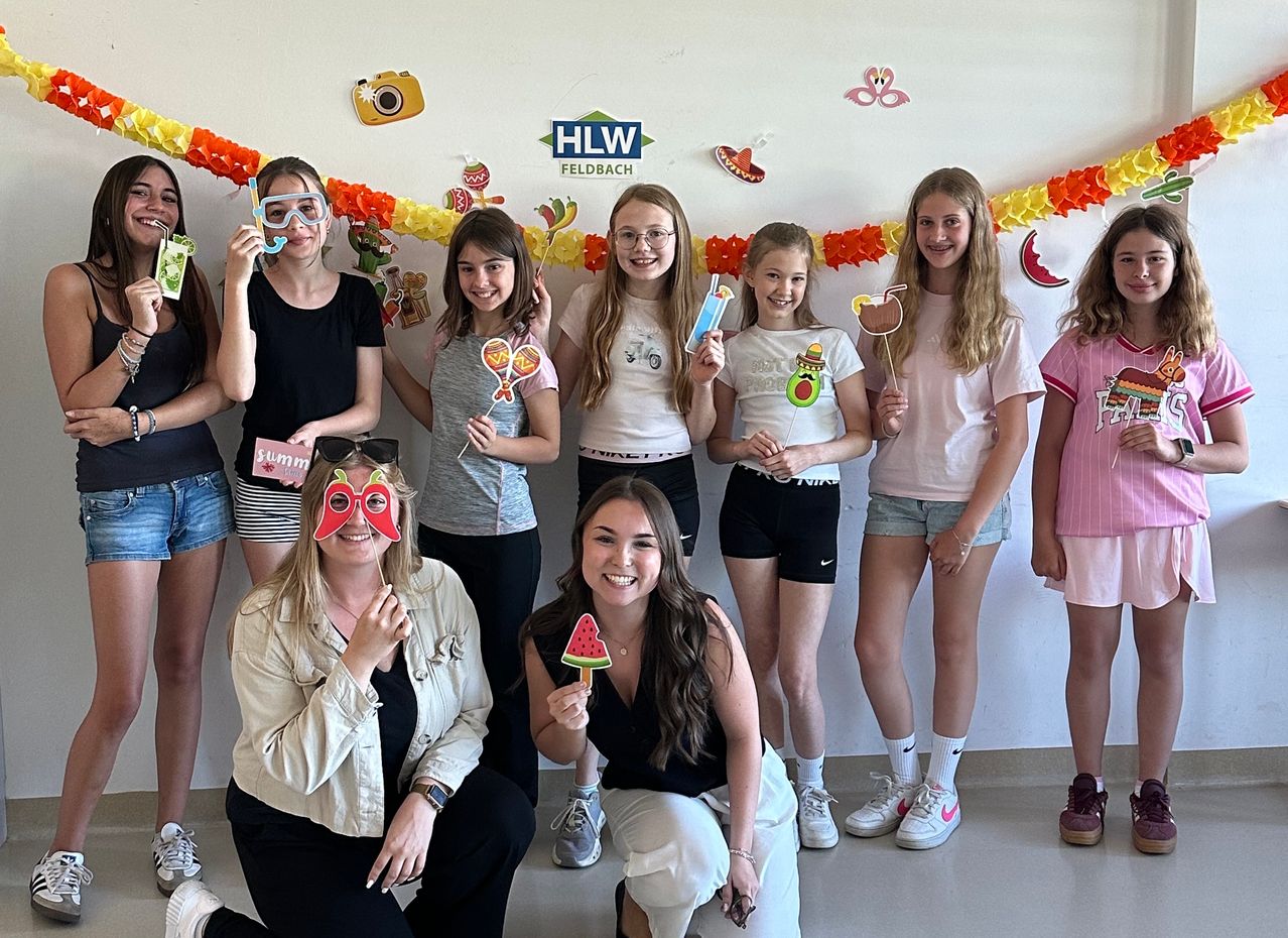 A group of girls and women pose for a photo. They hold props and wear casual attire. The wall behind them is decorated with colorful garlands.
