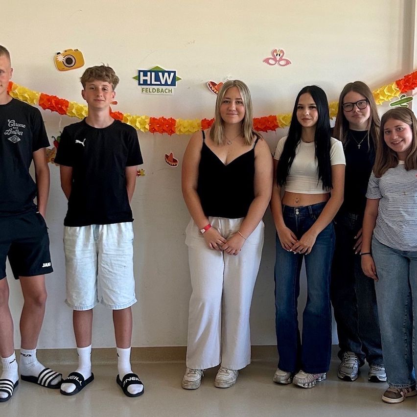 Six teenagers stand together for a photo in a room, with a white wall adorned with colorful paper garlands and a blue sign reading 'HLW Feldbach.' They are smiling and appear to be posing for a picture.