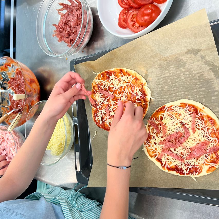 A person is making pizza with cheese and ham on a baking sheet on a table with various ingredients.