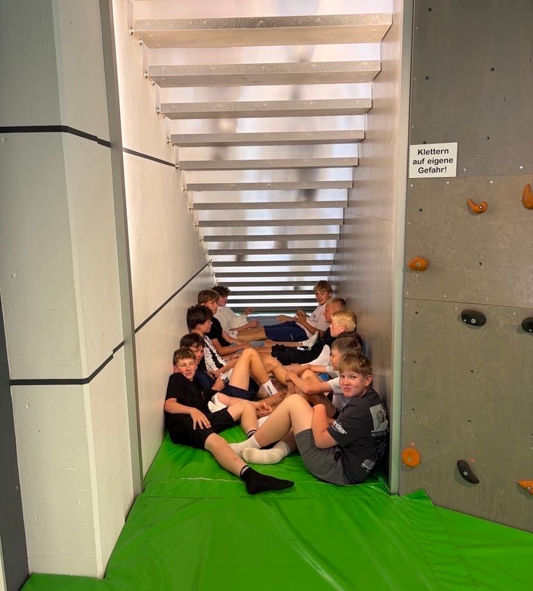 A group of young boys are sitting on a green mat in a small room. One boy is wearing a black shirt. There are stairs and a sign on the wall.