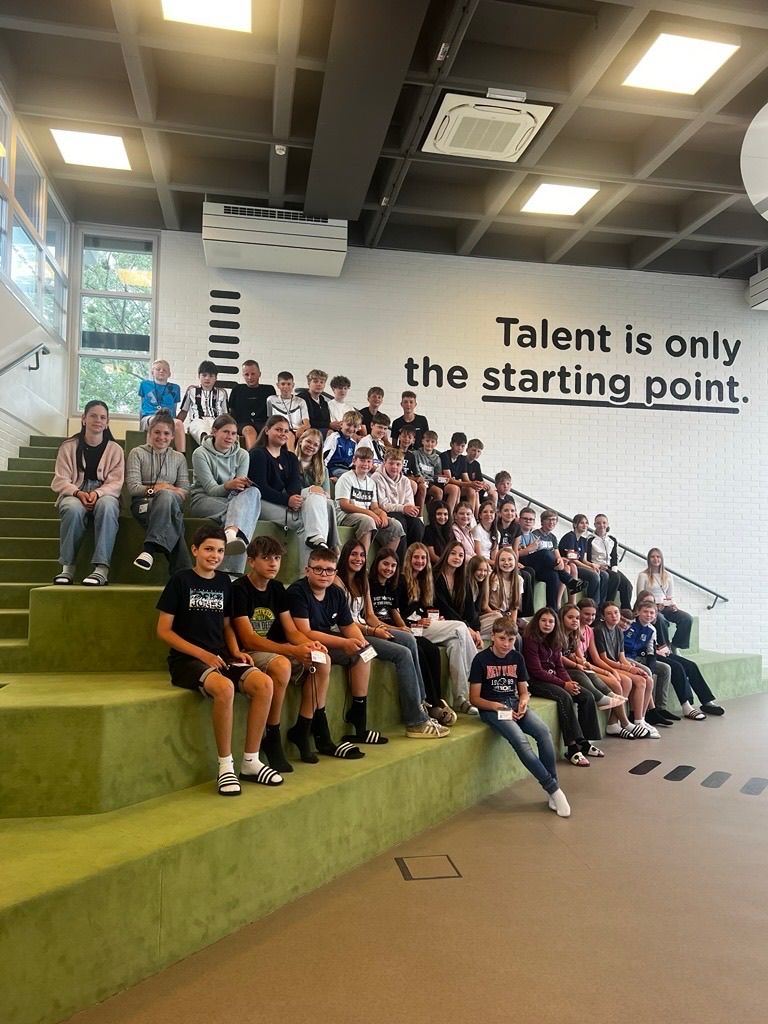 A group of students pose on green stairs with a motivational quote on the wall. They wear casual clothes and some have IDs. The room has white walls, glass windows, and an air vent.