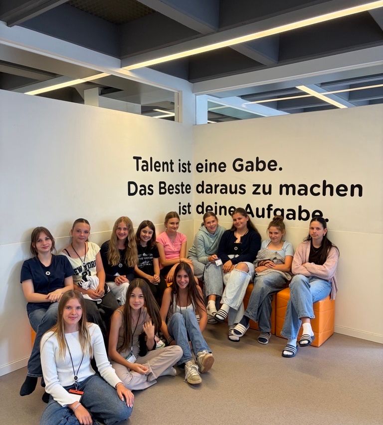 A group of young women sit together in an office. They are seated on orange chairs against a wall with a motivational quote.