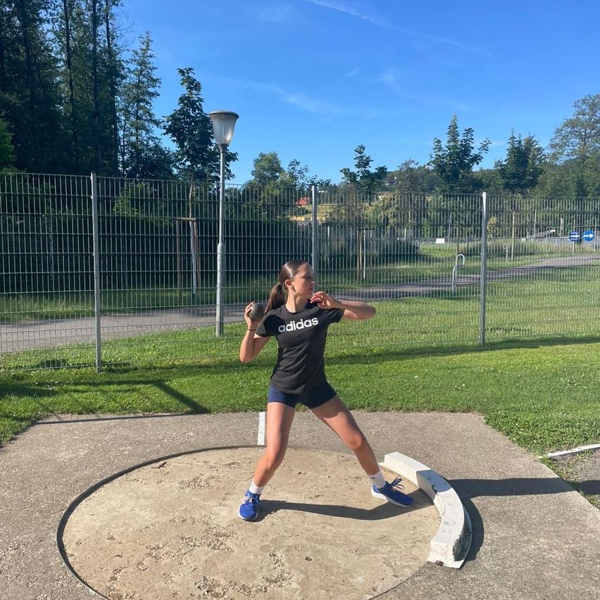 Eine Frau in Adidas-Sportkleidung wirft einen Diskus in einem Park. Hinter ihr steht ein Metallzaun, und Bäume und Gras sind sichtbar.