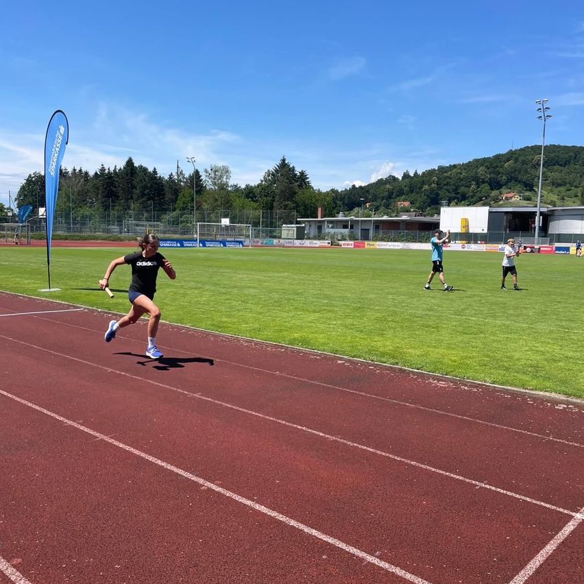 Eine Frau in schwarzer Adidas-Kleidung läuft auf einer Laufbahn. Im Hintergrund stehen zwei Personen auf einem Feld mit einem blauen Banner.