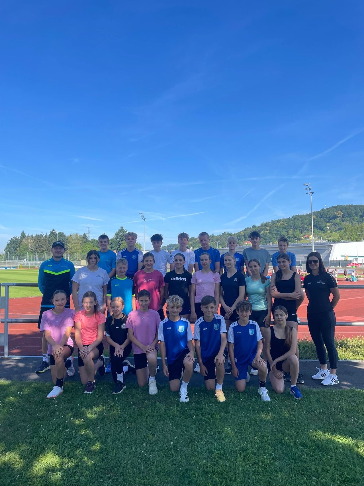 Eine Gruppe junger Athleten in Sportkleidung steht auf einem Sportplatz mit einem Metallzaun, lächelt und posiert für ein Foto. Sie sind von Grün umgeben und haben Berge in der Ferne.
