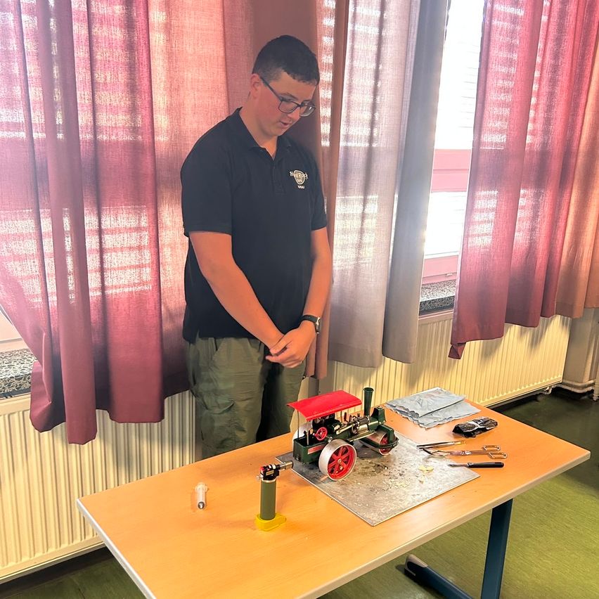 Ein Mann mit Brille steht in einem Raum, hält eine Taschenlampe und arbeitet an einer Spielzeug-Dampflokomotive auf einem Holztisch.