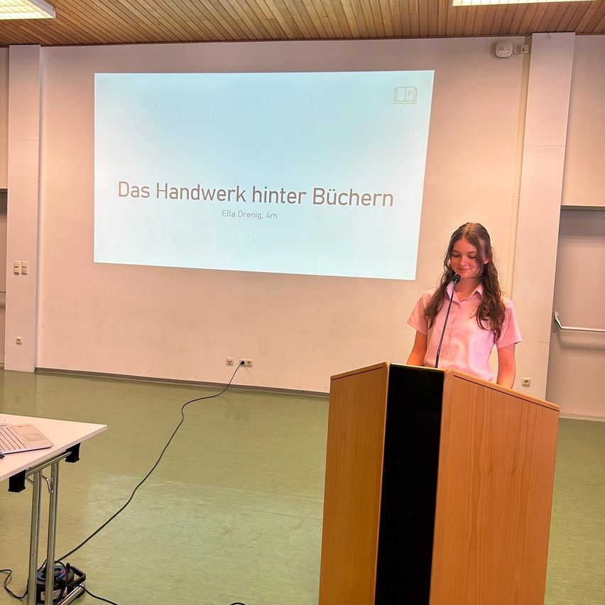 Ein junges Mädchen steht an einem Rednerpult und hält eine Präsentation. Ein Beamer hinter ihr zeigt 'Das Handwerk hinter Büchern'. Die Umgebung ist ein Klassenzimmer mit einem Tisch, einem Laptop und verschiedenen Steckdosen.