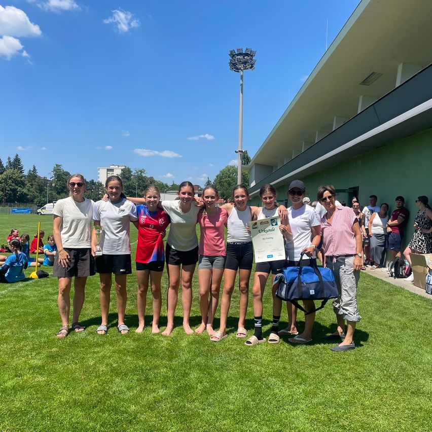 Eine Gruppe junger Frauen steht auf einem Rasenfeld, hält eine Trophäe und ein Zertifikat, trägt Sportbekleidung und Sneakers. Sie lächeln und posieren für ein Foto. Dahinter sind ein Gebäude, eine Straßenlaterne und eine Menschenmenge zu sehen.