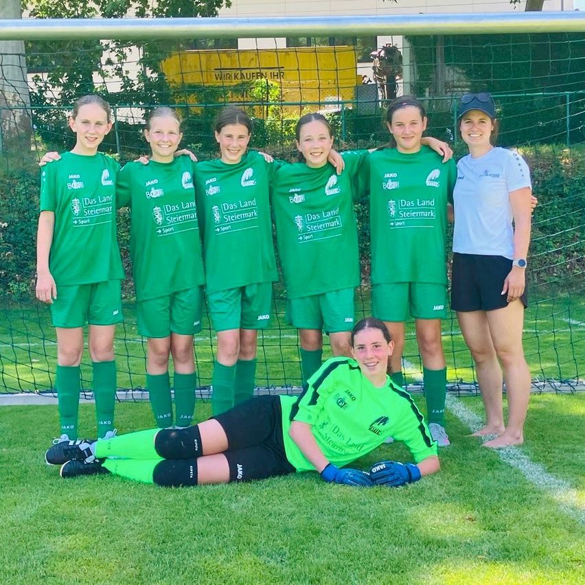 Eine Gruppe junger weiblicher Fußballerinnen in grünen Trikots mit 'Das Land Steiermark' darauf. Eine von ihnen liegt mit einem Lächeln auf dem Boden. Dahinter steht ein Tornetz und ein gelbes Banner mit Text.