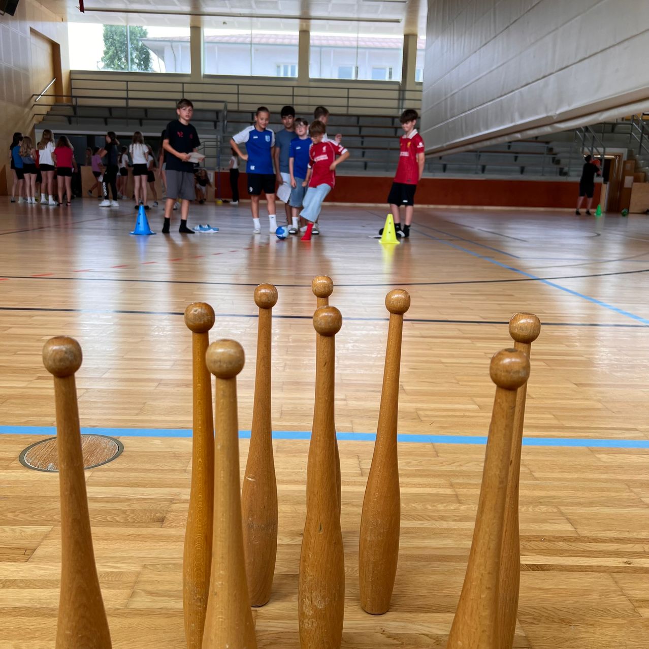 Eine Gruppe von Kindern spielt in einer Turnhalle mit Holzschlägern und Kegeln. Der Hallenboden hat blaue Markierungen.