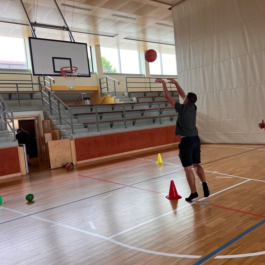 Ein Mann wirft einen Basketball in Richtung Korb auf einem Hallenplatz. Orangefarbene Kegel markieren die Grenzen des Spielfelds.