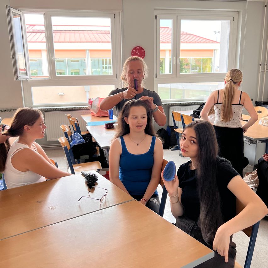 In einem Klassenzimmer frisiert eine Frau die Haare eines Mädchens mit einer Bürste. Ein anderes Mädchen beobachtet, während ein anderes am Tisch sitzt. Zwei weitere Frauen sind im Hintergrund.