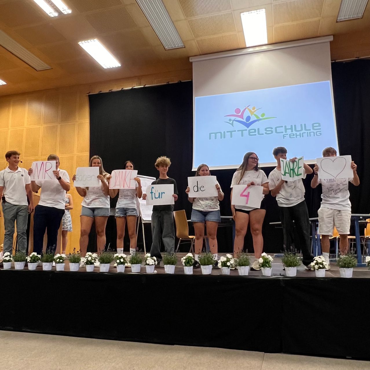 Students stand on stage holding signs with names and numbers, in front of a presentation screen. They appear to be participating in an event or competition.