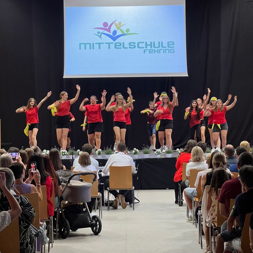 A group of people on stage, wearing red shirts and black shorts, perform in front of an audience. A large screen in the background displays the 'Mittelschule Ferching' logo.