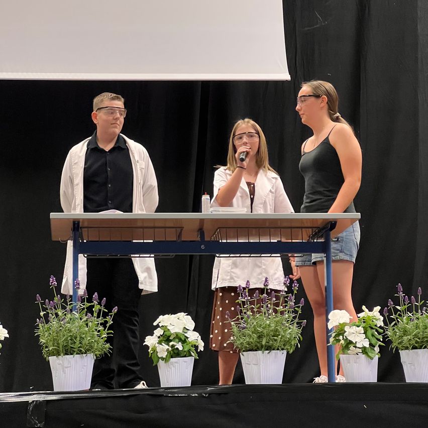 Three individuals stand on a stage, two women and a man, with plants in white pots. The man wears glasses and a lab coat. One woman speaks into a microphone while the other watches.