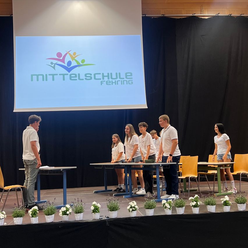 A group of students stands on a stage in front of tables, with a screen displaying the Mittelschule Fehring logo. Two men and several girls are present.