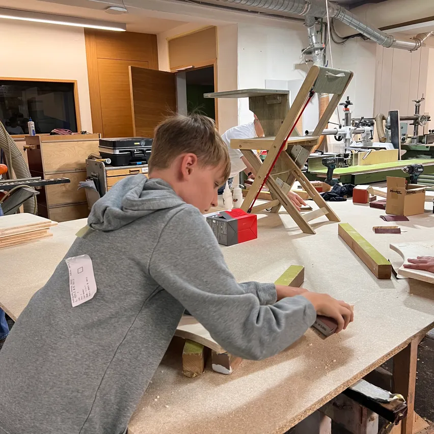 Ein junger Junge in einer grauen Kapuzenjacke arbeitet in einer Werkstatt an einem Holzprojekt. Er schleift ein Stück Holz auf einem Tisch. Verschiedene Werkzeuge und Materialien sind um ihn herum.