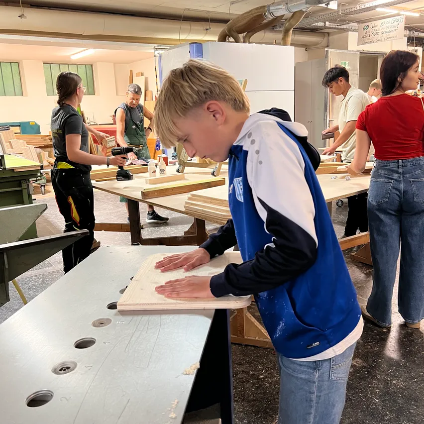In einer Werkstatt arbeiten mehrere Personen an Holzbrettern, ein Junge in einem blau-weißen Jackett konzentriert sich auf seine Aufgabe.