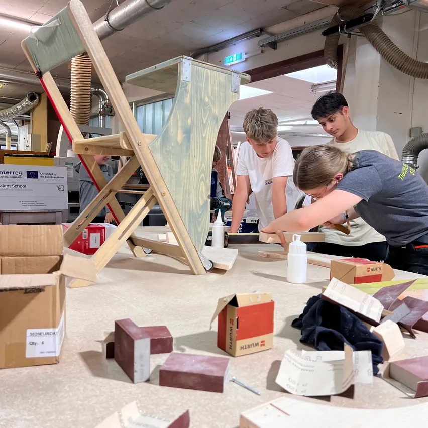 Drei Personen arbeiten in einer Werkstatt an einem Holzstuhl. Einer trägt Klebstoff auf, ein anderer hält ein Stück Holz und der dritte beobachtet. Verschiedene Materialien und Werkzeuge sind herumgestreut.