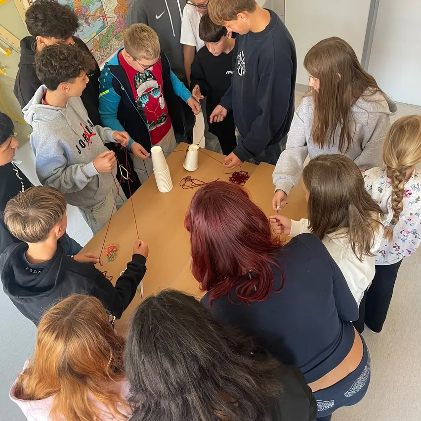 Eine Gruppe von Kindern und einigen Erwachsenen versammelt sich um einen Tisch und beschäftigt sich mit einer Aktivität mit Schnüren und Bechern.