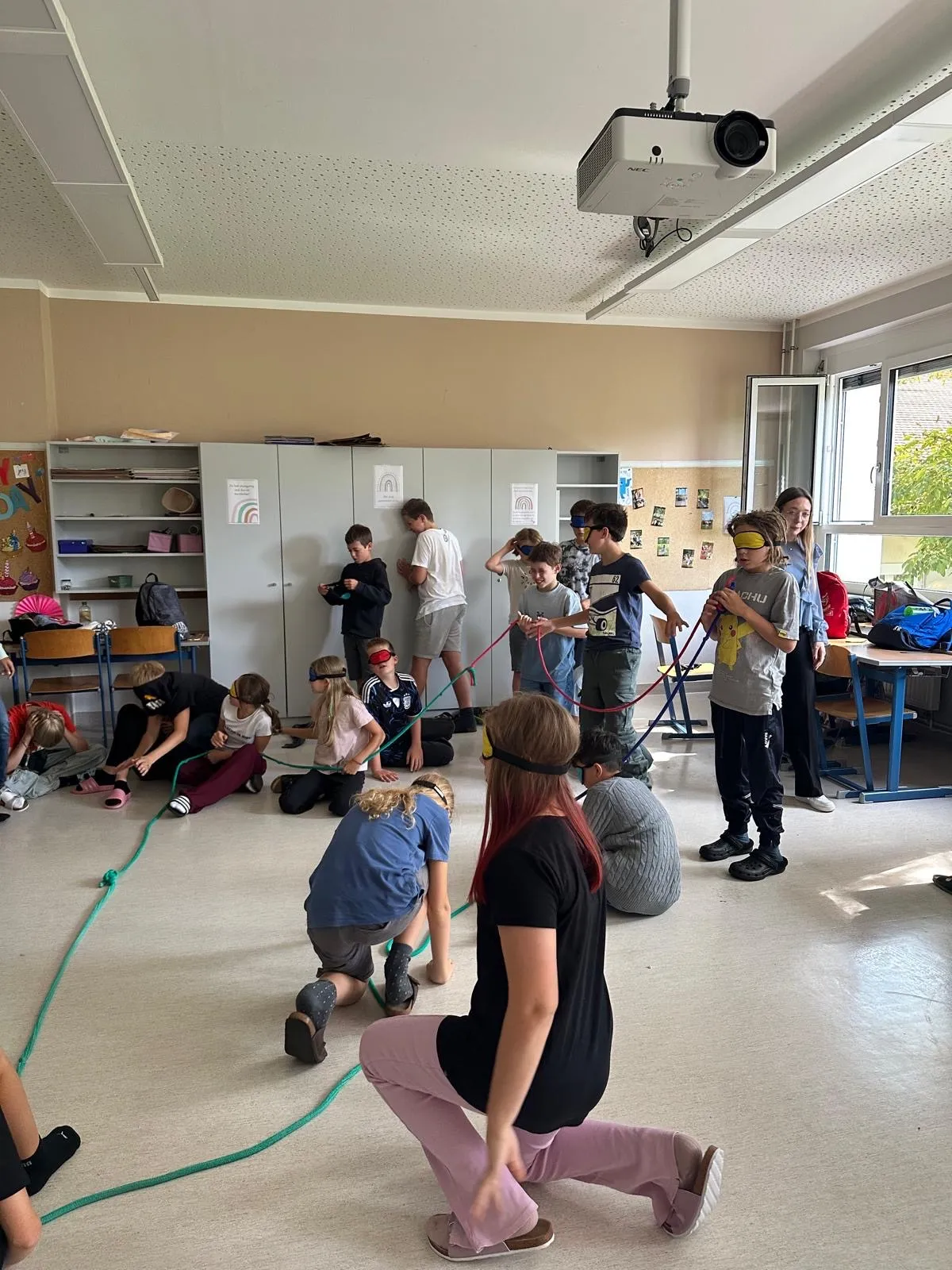 Eine Gruppe von Kindern und Erwachsenen nimmt in einem Klassenzimmer an einer Blindenaktivität teil. Sie halten sich an einem grünen Seil fest und führen sich gegenseitig durch den Raum.