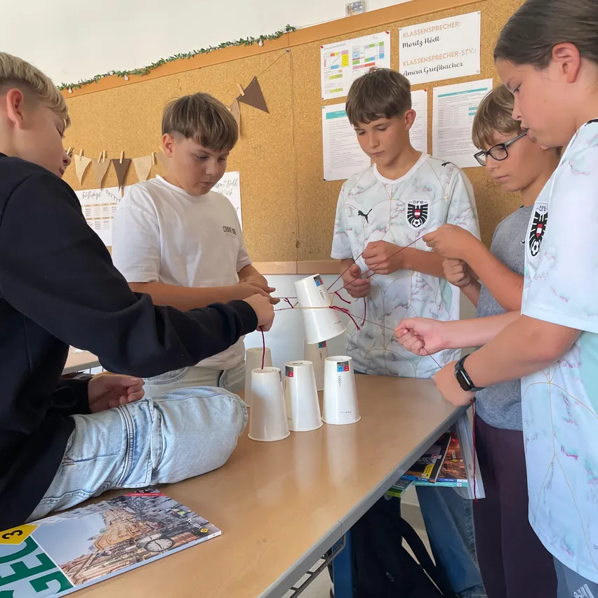Fünf Schüler sitzen um einen Tisch herum und arbeiten an einem Projekt mit Bechern und Drähten. Sie scheinen konzentriert zu sein.