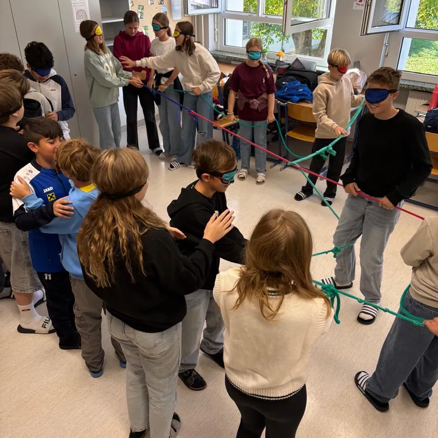 Eine Gruppe von Kindern mit verbundenen Augen hält sich an den Händen und bildet einen Kreis in einem Raum.