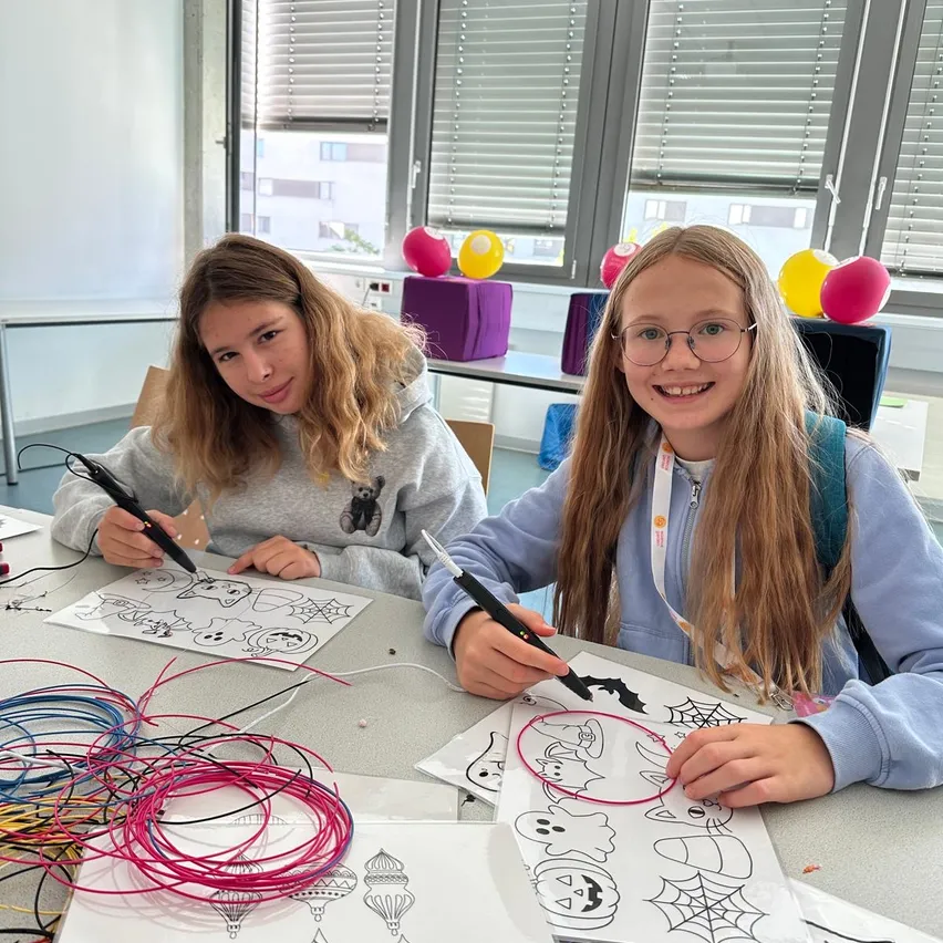 Zwei junge Mädchen sitzen an einem Tisch und halten jeweils einen Stift in der Hand und zeichnen auf einem Blatt Papier. Sie sind von bunten Drähten und Luftballons umgeben.
