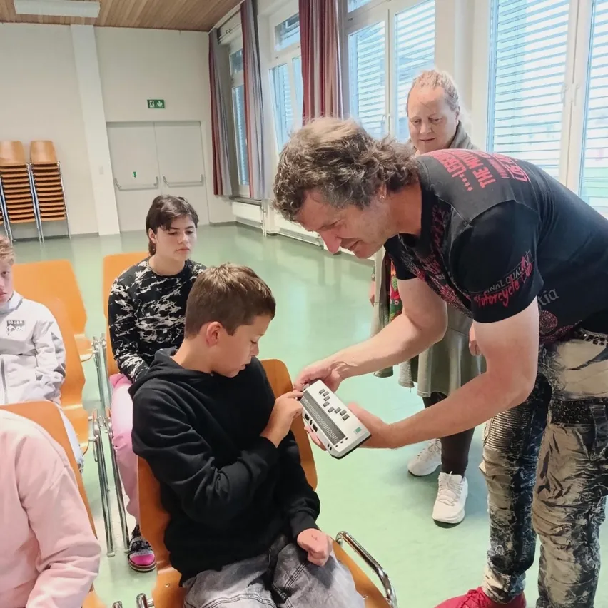 Ein Mann zeigt einem jungen Jungen in einem Stuhl ein Gerät, während andere in einem Klassenzimmer zuschauen.