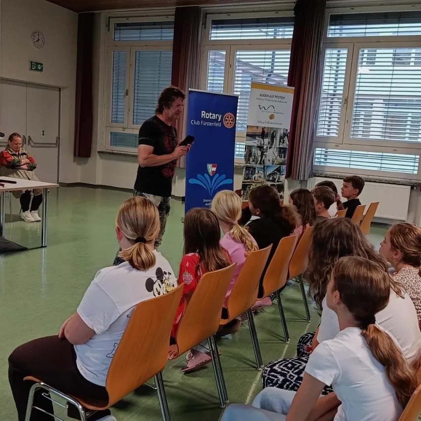Ein Mann steht vor einer Gruppe von Kindern und hält ein Mikrofon. Die Kinder sitzen in Stühlen und schauen ihm zu. Hinter ihm steht ein Banner mit der Aufschrift 'Rotary Club Furstenfeld.'