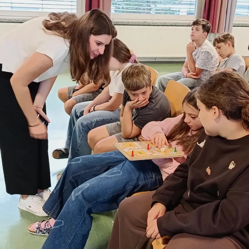 Eine Frau in einem weißen Shirt beugt sich vor, um Kinder beim Spielen eines Brettspiels zu beobachten. Die Kinder sitzen auf Stühlen, einige mit den Händen am Kinn.