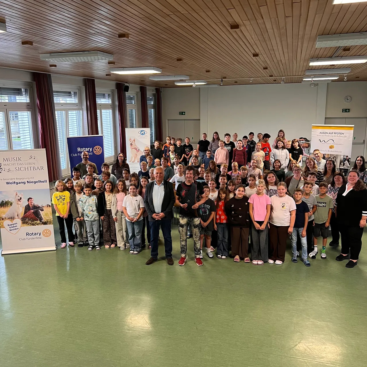 Eine große Gruppe von Kindern und Erwachsenen versammelt sich in einem Raum und posiert für ein Foto. Banner sind auf der linken und rechten Seite angebracht.