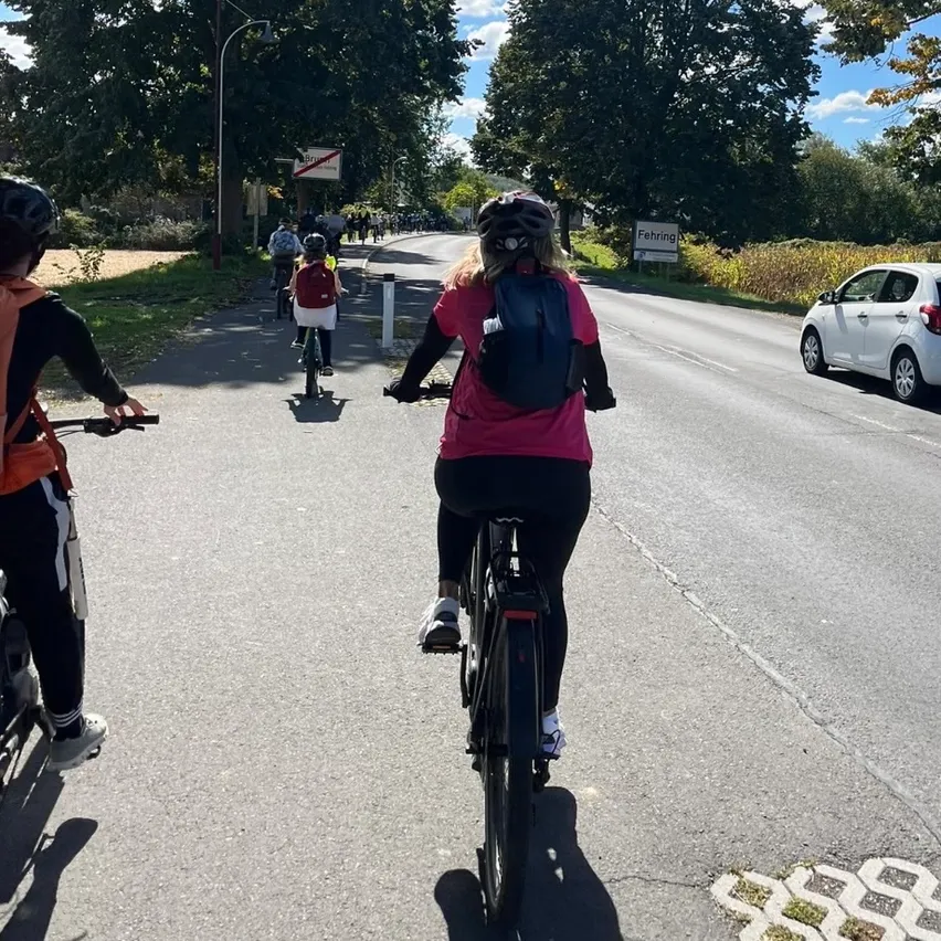 Eine Gruppe von Radfahrern fährt auf einer Straße. Die Radfahrer tragen Helme und tragen Rucksäcke. Ein weißes Auto fährt hinter ihnen auf der Straße.