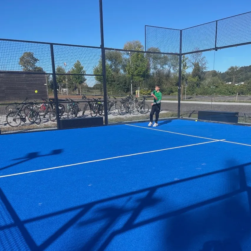 Ein Tennisspieler steht auf einem blauen Platz, möglicherweise bereit zum Aufschlag. Der Platz ist von einem Netzzaun umgeben, und mehrere Fahrräder sind draußen geparkt.