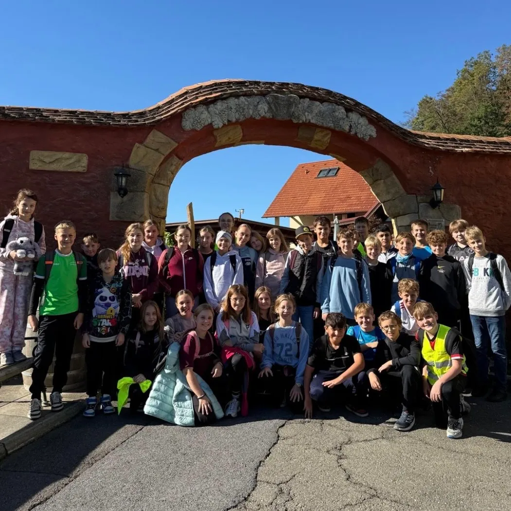 Eine Gruppe von Kindern und Jugendlichen posiert für ein Foto vor einem Bogen. Der Bogen ist aus Stein und hat ein geschwungenes Design. Die Kinder tragen verschiedene Kleidungsstücke und einige von ihnen tragen Rucksäcke. Dahinter befindet sich ein Gebäude mit einem roten Dach.