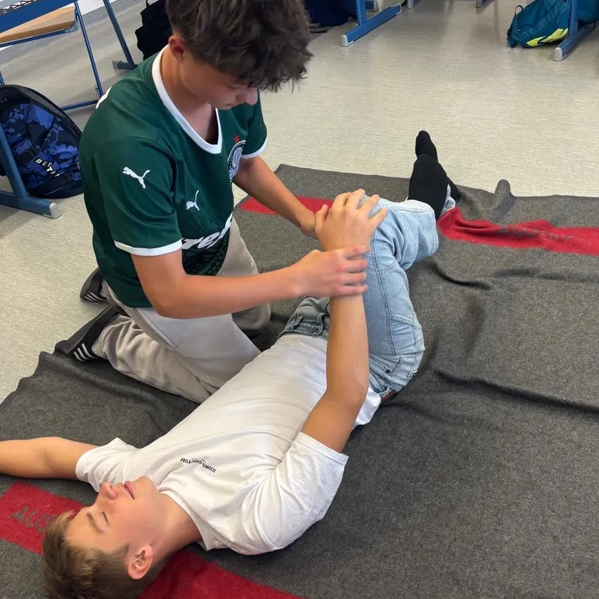 Ein junger Mann liegt auf einer Decke auf dem Boden, ein anderer Mann kniet neben ihm und führt möglicherweise eine physiotherapeutische Übung durch.