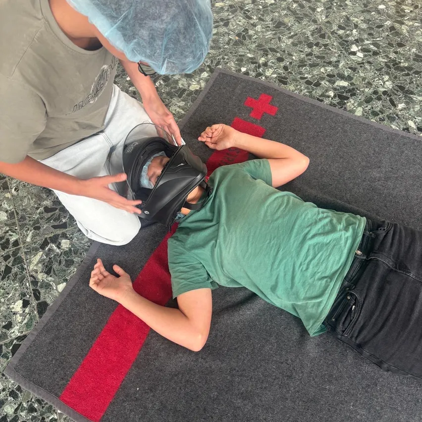 Eine Person mit einem blauen Haarnetz und einem grauen T-Shirt kniet auf dem Boden und hält einen Helm über eine andere Person, die auf einem grauen Teppich mit einem roten Kreuzsymbol liegt.