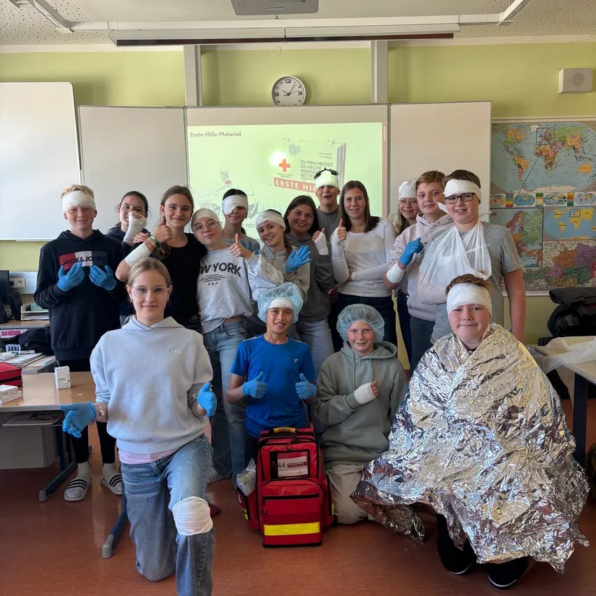 Eine Gruppe von Schülern in einem Klassenzimmer nimmt an einer Erste-Hilfe-Simulation teil. Sie tragen Augenbinden, Verbände und Handschuhe, und einige sind mit silbernen Decken bedeckt.