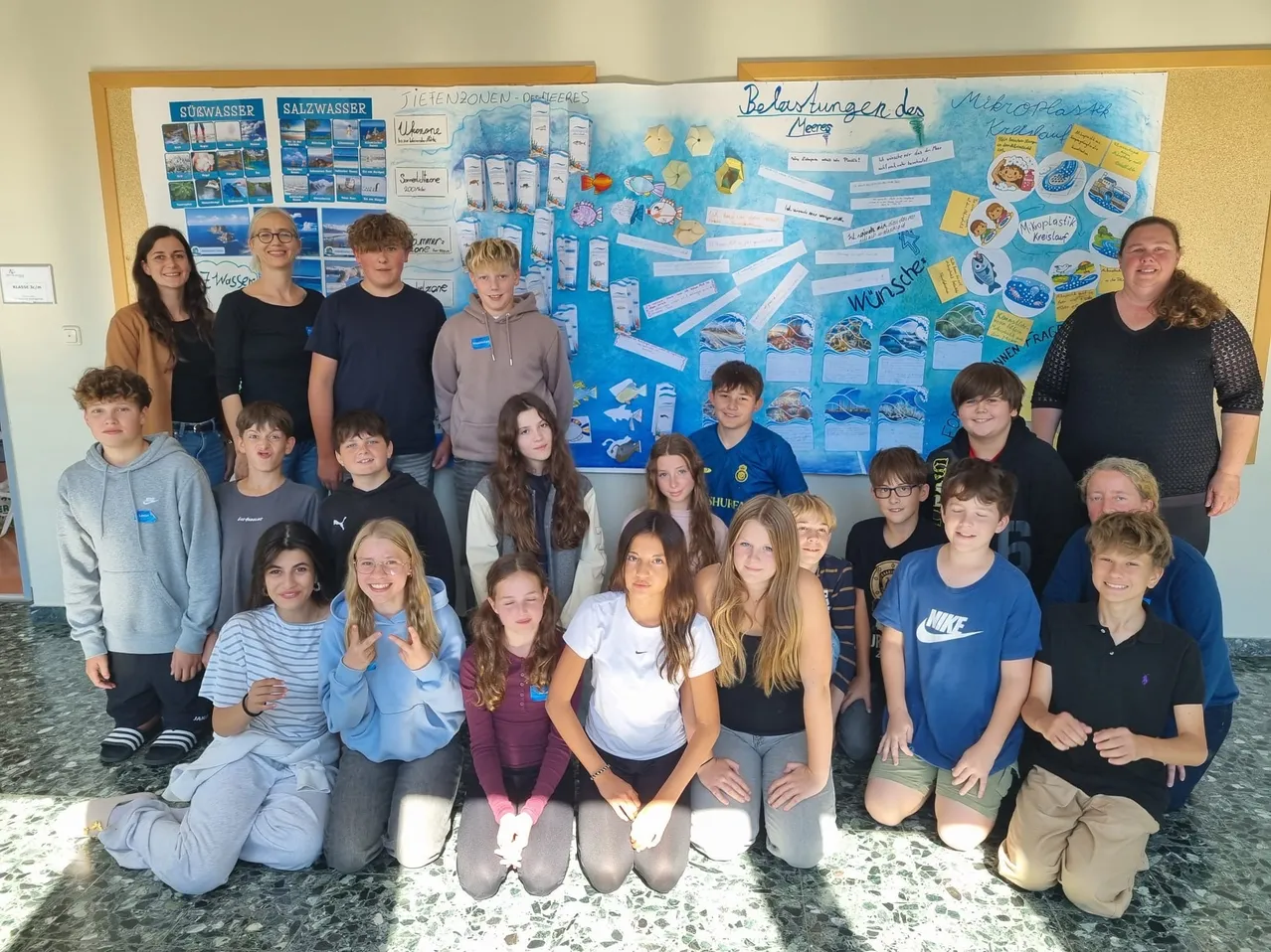 Eine Gruppe von Schülern und Lehrern posiert für ein Foto vor einem großen Poster. Das Poster hat einen blauen Hintergrund mit verschiedenen Bildern und Text.