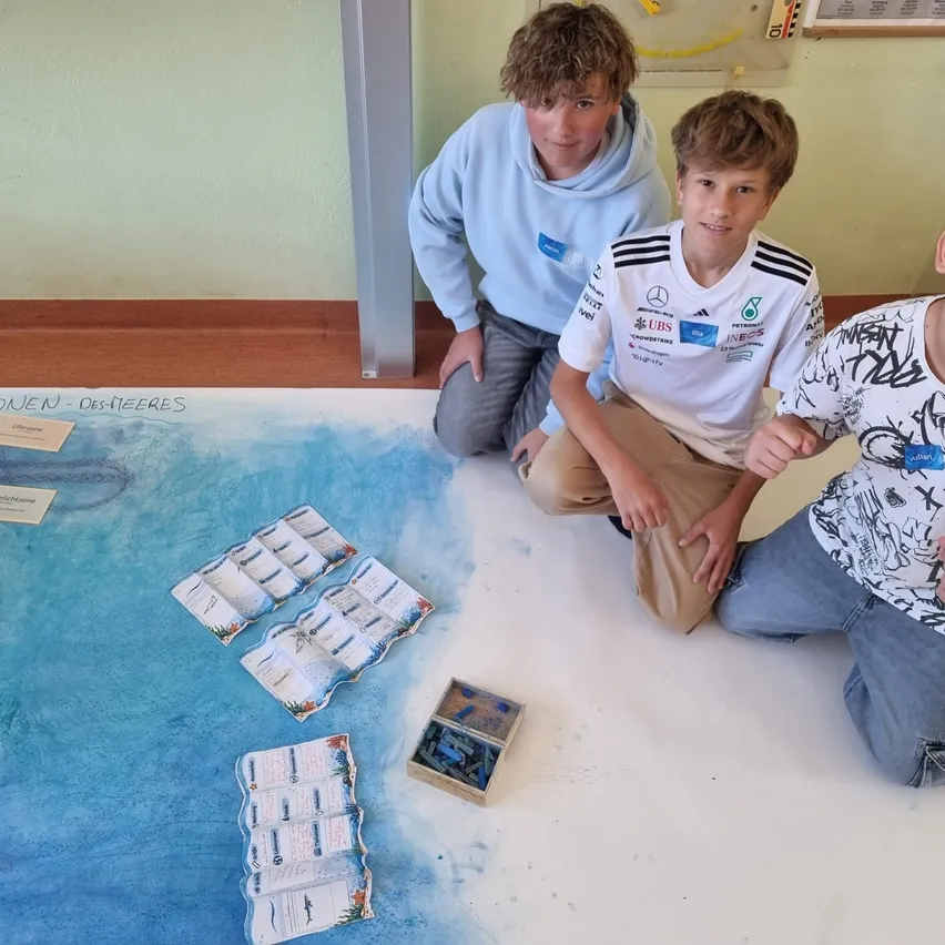 Vier Jungen knien auf dem Boden, lächeln und posieren für ein Foto. Sie tragen verschiedenfarbige Hemden. Es gibt ein großes blau-weißes Poster auf dem Boden mit einigen Karten und einer Schachtel.