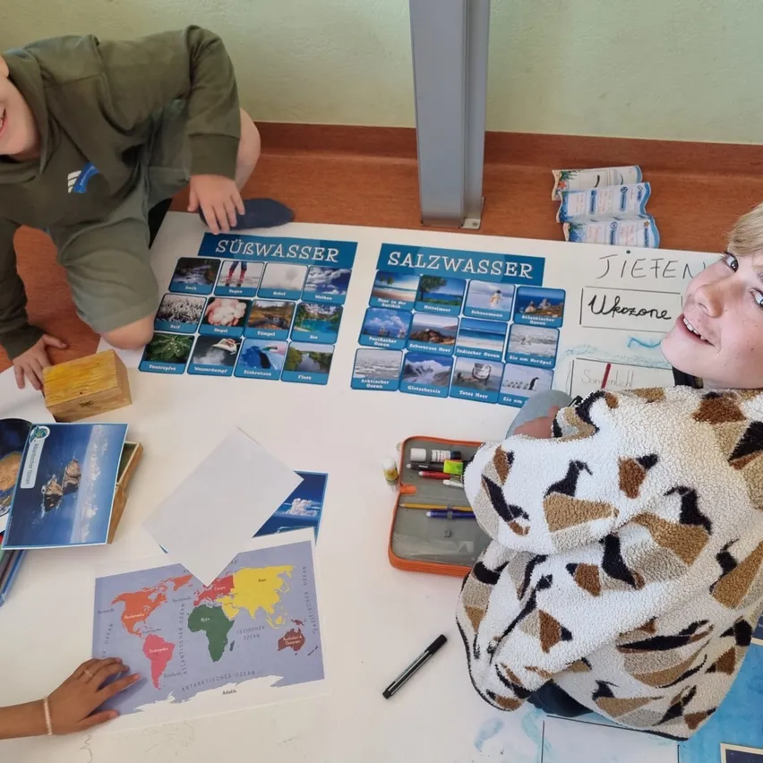 Zwei Kinder sitzen auf dem Boden um einen Tisch mit einer Weltkarte, Büchern und einem Stift herum. Ein Kind lächelt und schaut in die Kamera.