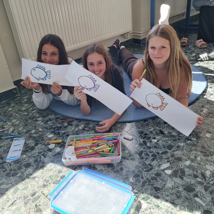 Drei junge Mädchen liegen auf dem Boden, lächeln und halten Papier mit Fischzeichnungen hoch. Sie sind von farbigen Stiften und anderen Zeichenutensilien umgeben.