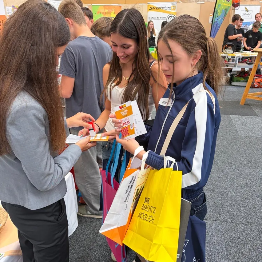 Eine Gruppe junger Leute steht in einem Raum, wahrscheinlich auf einer Messe. Eine Frau gibt einer anderen Frau einen Pass.