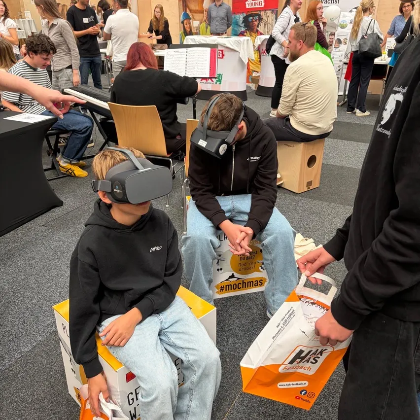 Zwei junge Jungen sitzen auf Kisten und tragen VR-Brillen, mit einem Mann, der einen Beutel mit Essen hinter ihnen hält.